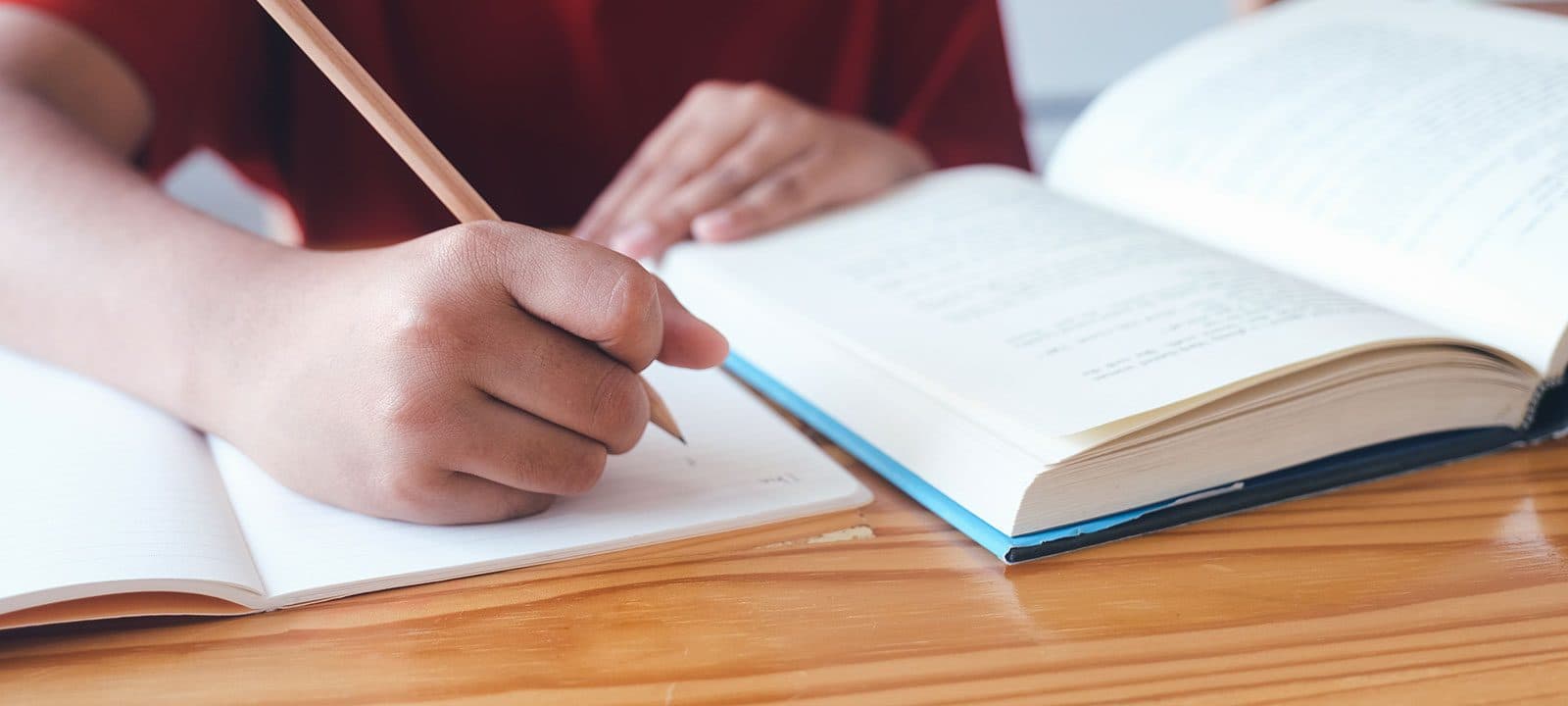 Student writing in a notebook with a pencil beside an open book.